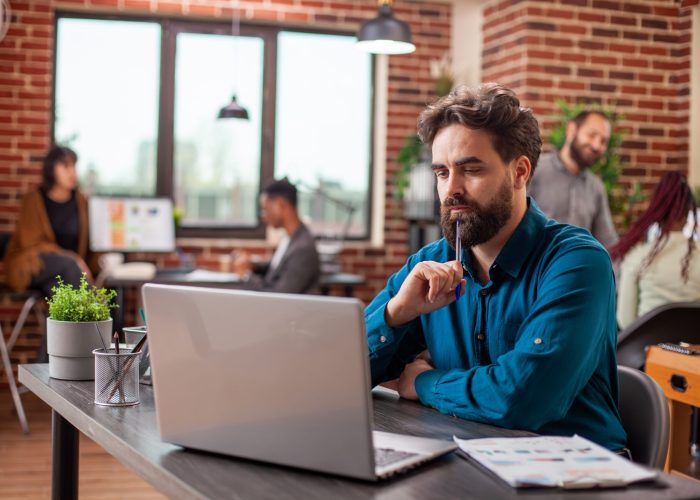 Male researcher studying documents with pen in hand, focused on project development at desk. Caucasian employee seated in brick wall office, tapping his chin and reviewing business plans on laptop.